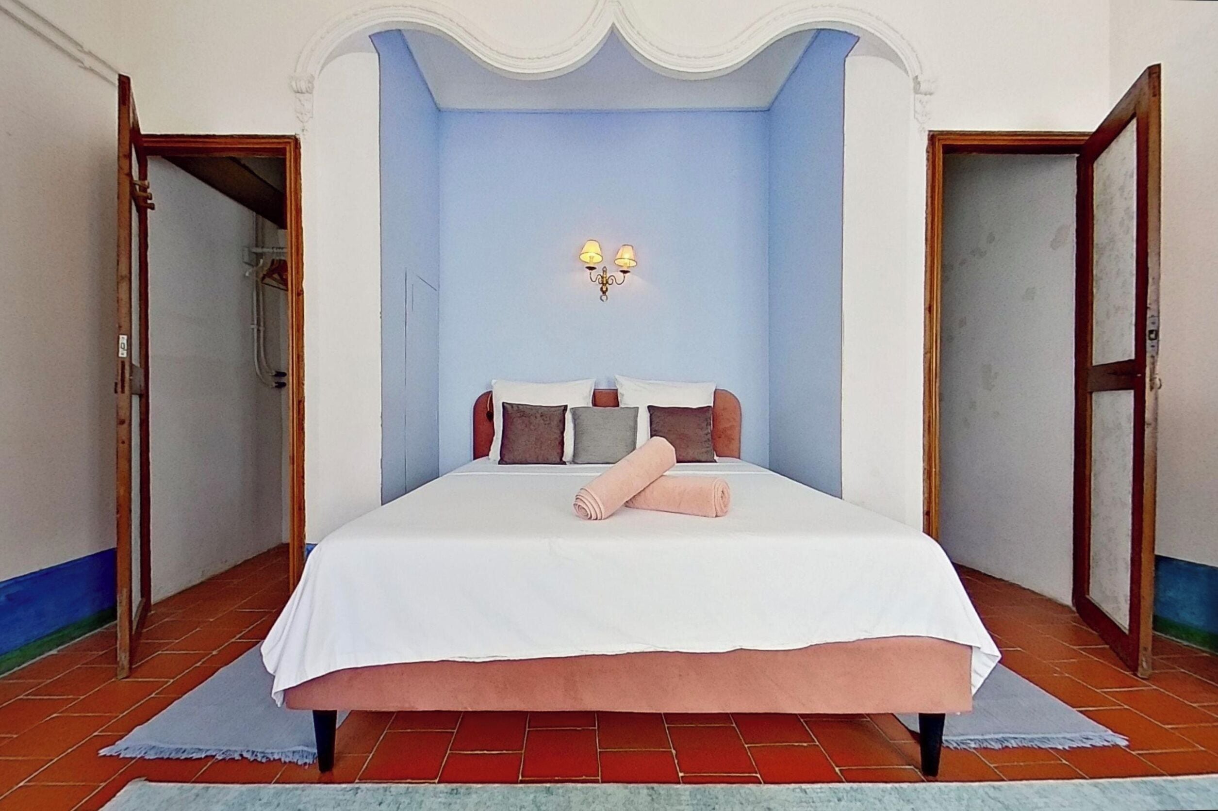 Un dormitorio con una cama grande, una mesita de noche, una lámpara de pared y una puerta que da a otra habitación.