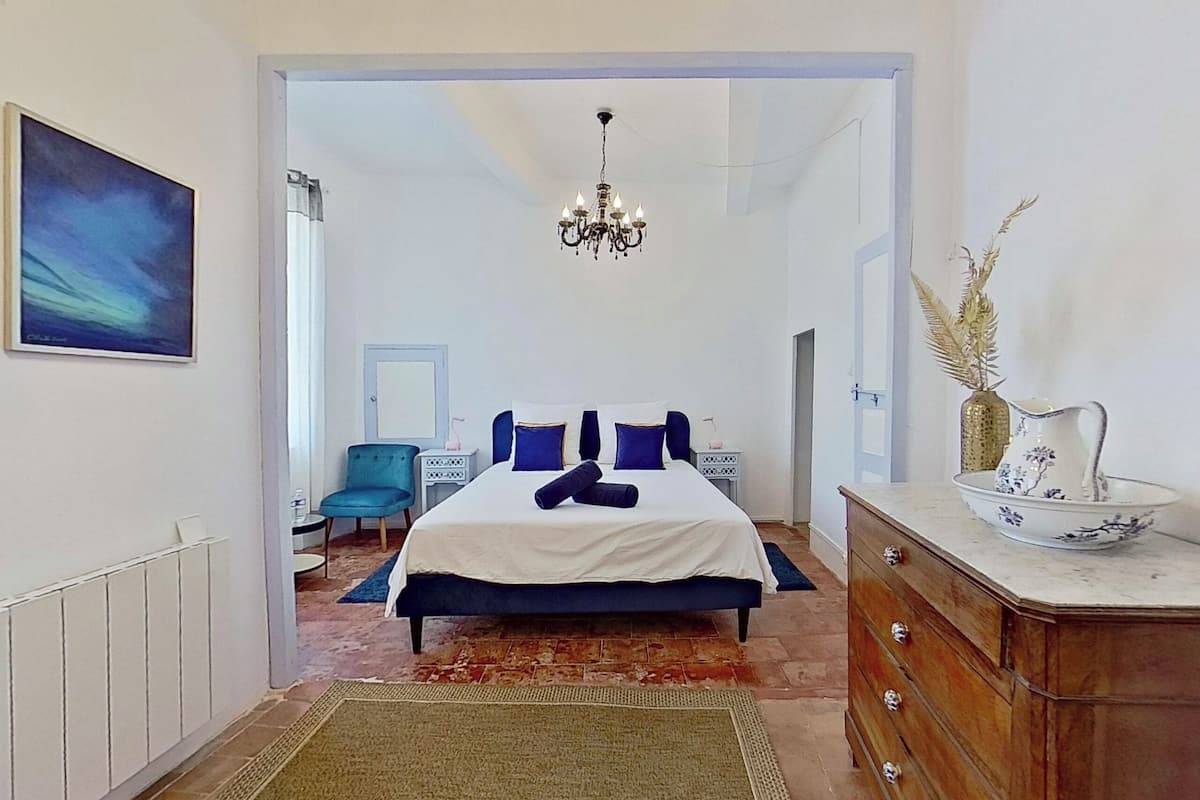 A bedroom with a bed, a dresser, a chair, a chandelier, and a painting on the wall.