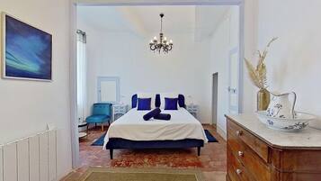 A bedroom with a bed, a dresser, a chair, a chandelier, and a painting on the wall.