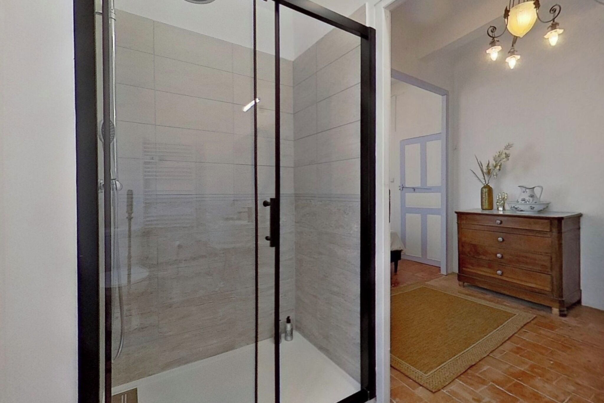 Une douche fermée par des parois de verre, un mur recouvert de carreaux, une commode en bois ornée d’un vase et d’une carafe, et un luminaire suspendu.