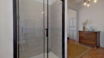 Ducha con cerramiento de vidrio, pared con azulejos, cómoda de madera con un florero y una jarra, y lámpara colgante.