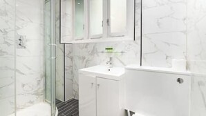 Une salle de bain moderne avec une douche à parois de verre, des murs en marbre blanc et un lavabo blanc agrémenté d’un distributeur de savon vert.