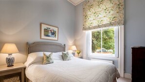 A bedroom with a large bed, two bedside tables with lamps, a framed picture on the wall, and a window with floral curtains.