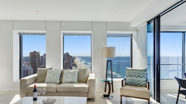 A modern living room with a sofa, a coffee table, and a chair. There are large windows offering a view of the ocean and cityscape.