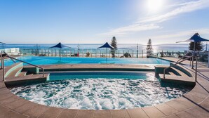 A pool area with a view of the ocean, a hot tub, and umbrellas providing shade.