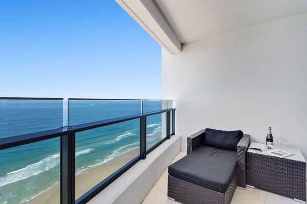 Un balcon avec vue sur l’océan, un canapé en osier, une petite table avec une bouteille et des verres, et un mur blanc.