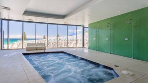 A modern indoor pool area with a hot tub, large windows offering a view of the ocean, and a green wall with a shower area.