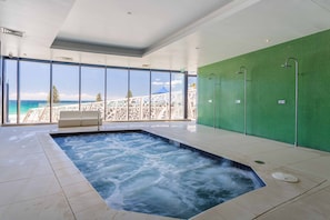 A modern indoor pool area with a hot tub, large windows offering a view of the ocean, and a green wall with a shower area.