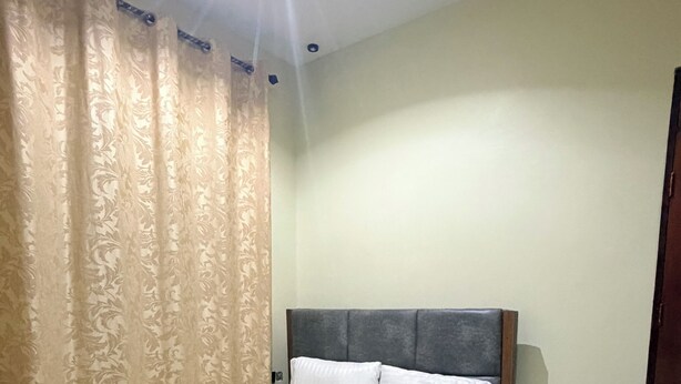 A single bed with a wooden frame and a grey headboard, a white mattress, and two white pillows. The room has a tiled floor and beige walls.