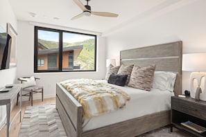 Relax in this serene bedroom with a large bed, desk, and nightstand, featuring a ceiling fan and a window with a view.