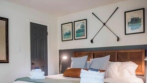 Un lit bien fait, avec du linge de lit blanc et un plaid turquoise, une tête de lit en bois et des œuvres d’art encadrées accrochées au mur.