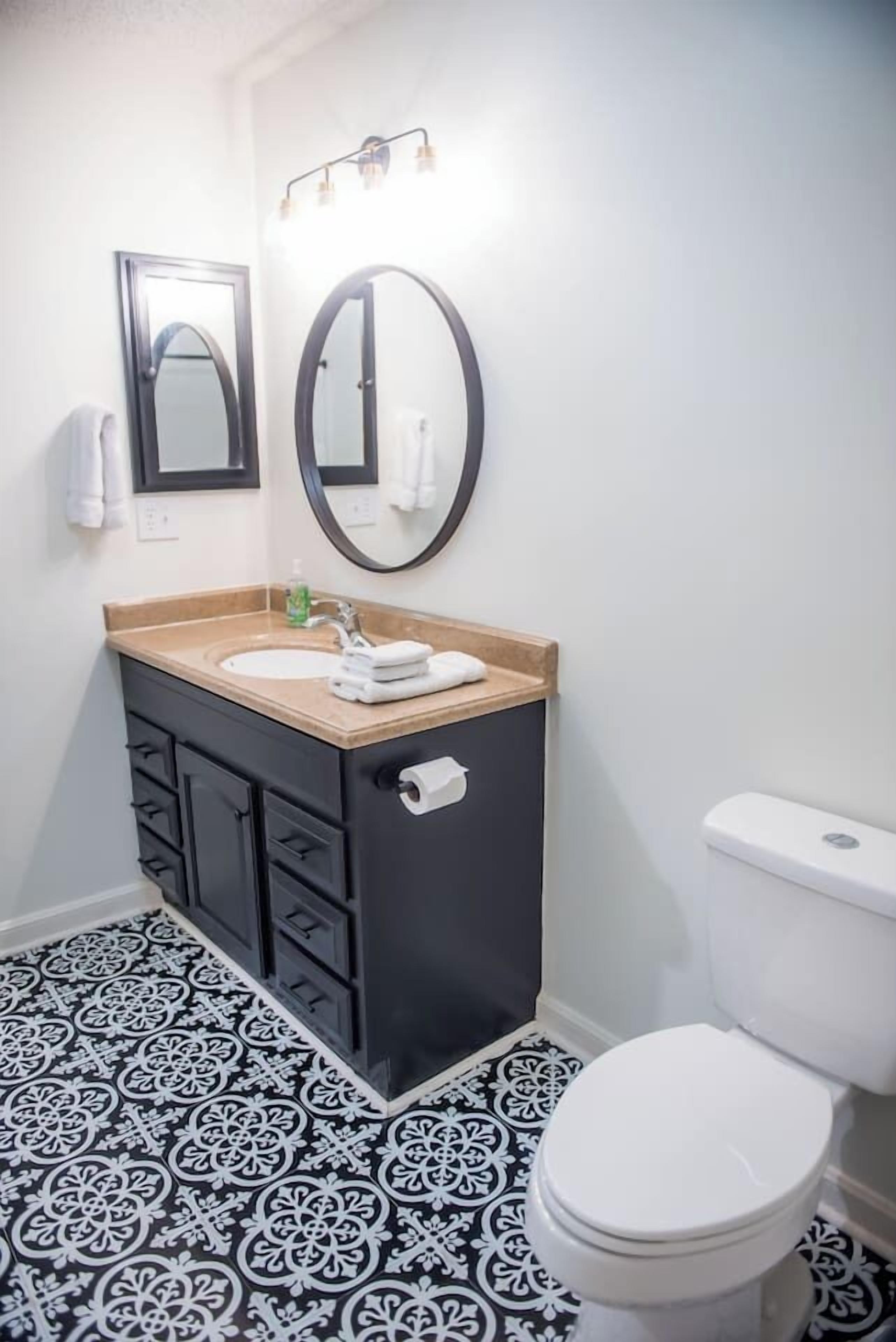 A bathroom with a black and white patterned floor, a black vanity with a beige countertop, a white toilet, and two mirrors with light fixtures above them.