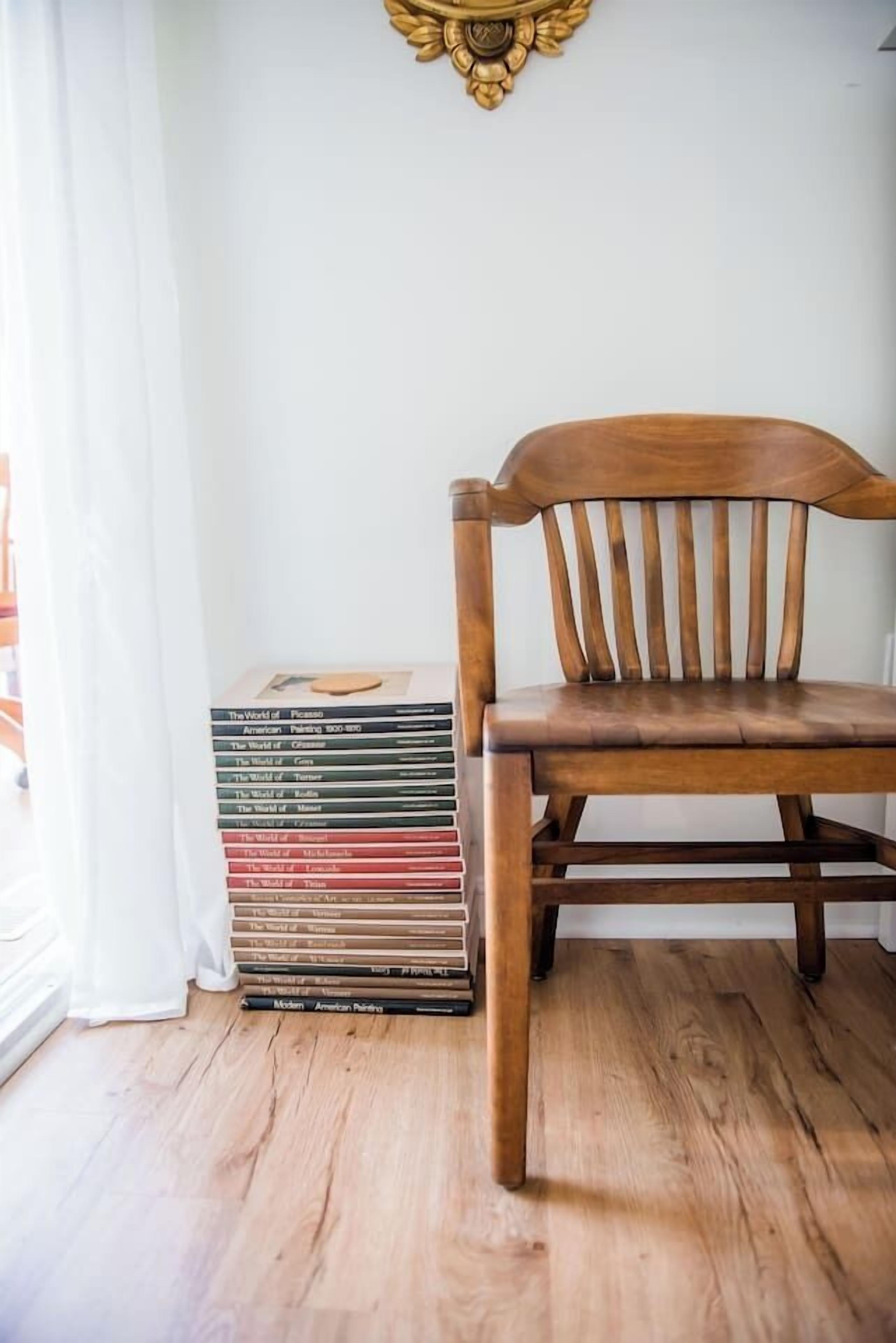 Una sedia di legno in una stanza con una pila di libri sul pavimento e un elemento decorativo sopra la parete.