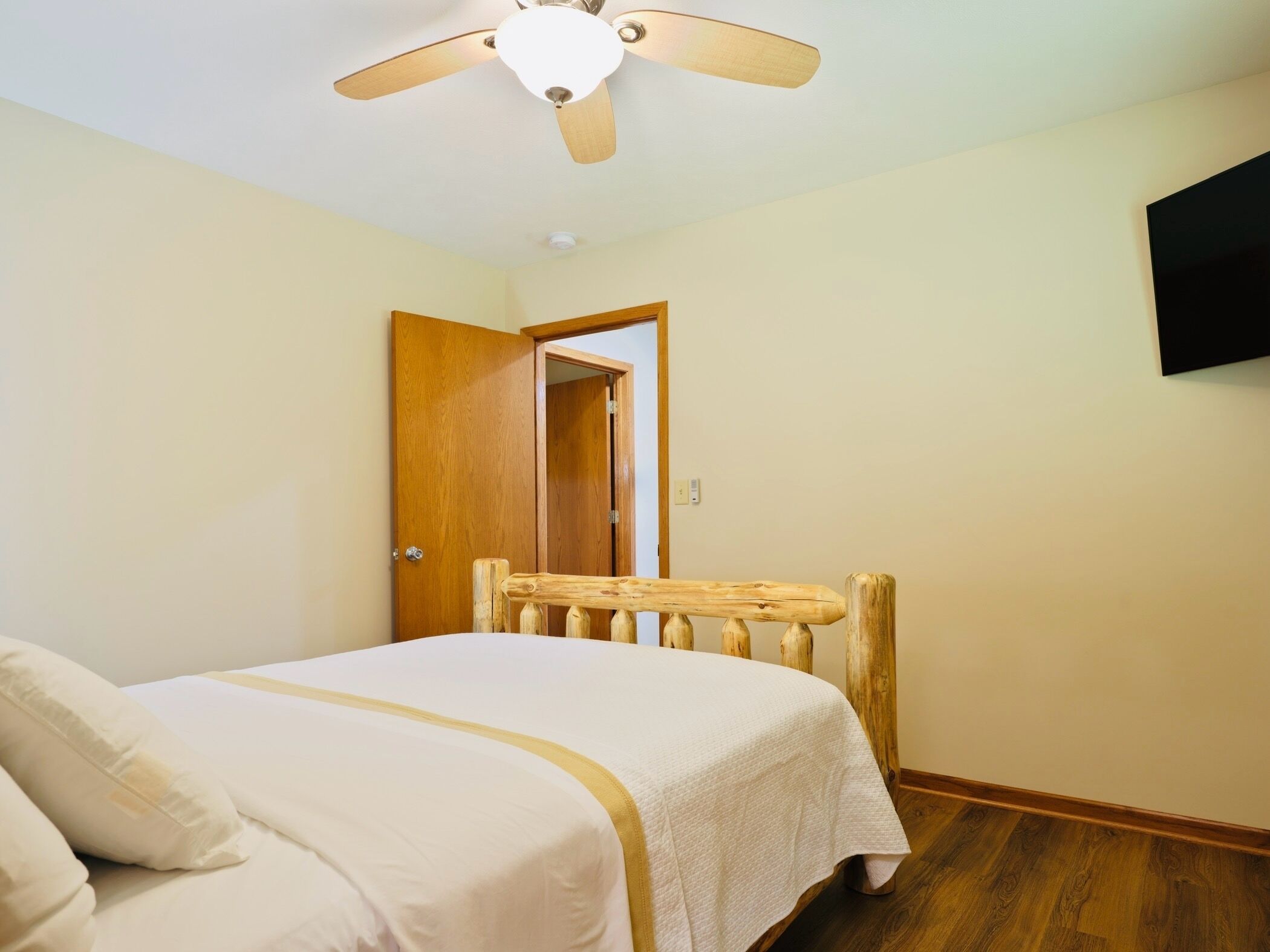 A bedroom with a bed, wooden headboard, ceiling fan, and a TV mounted on the wall.