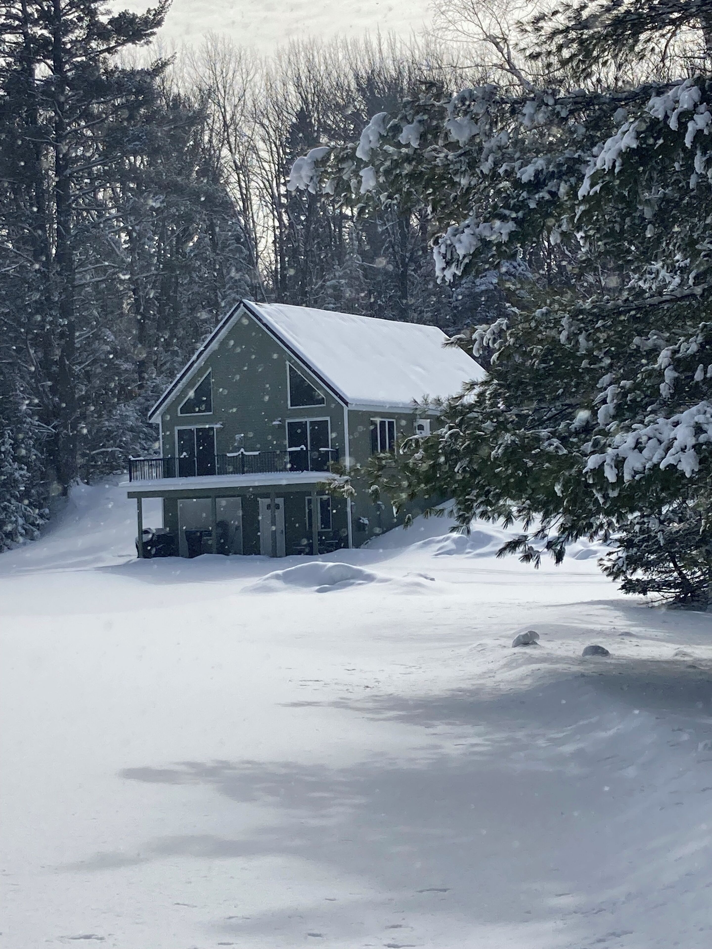 A two-story house with a snow-covered roof and a balcony, surrounded by trees and a snowy landscape.