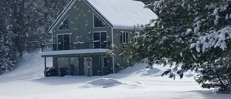 A two-story house with a snow-covered roof and a balcony, surrounded by trees and a snowy landscape.