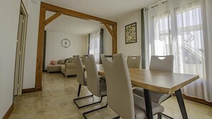 A dining area with a wooden table and chairs, a sofa in the background, and a view of a road through the window. - Maison des 3 Lacs - CHAVANGES (CHAVANGES)