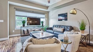 Ein Wohnzimmer mit Deckenventilator, einem großen Fenster mit Jalousien, einem Ledersofa, einem weißen Sessel, einem Couchtisch und einer Stehlampe.