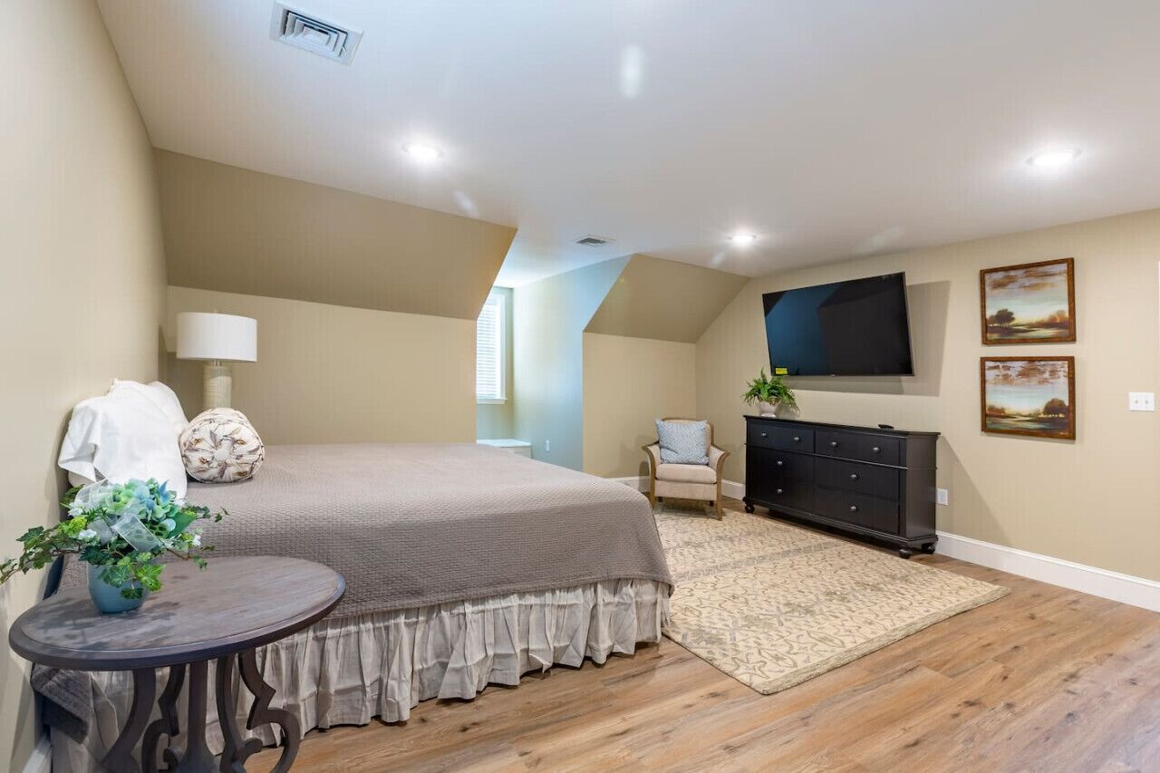 A bedroom with a large bed, a dresser, a TV mounted on the wall, and a small table with a plant.