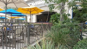 An outdoor seating area with tables and chairs, covered by umbrellas, and surrounded by greenery.