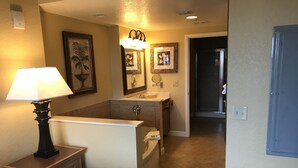 A hotel room with a bed, a nightstand with a lamp, a bathroom with a sink and mirror, and a hallway leading to another room.