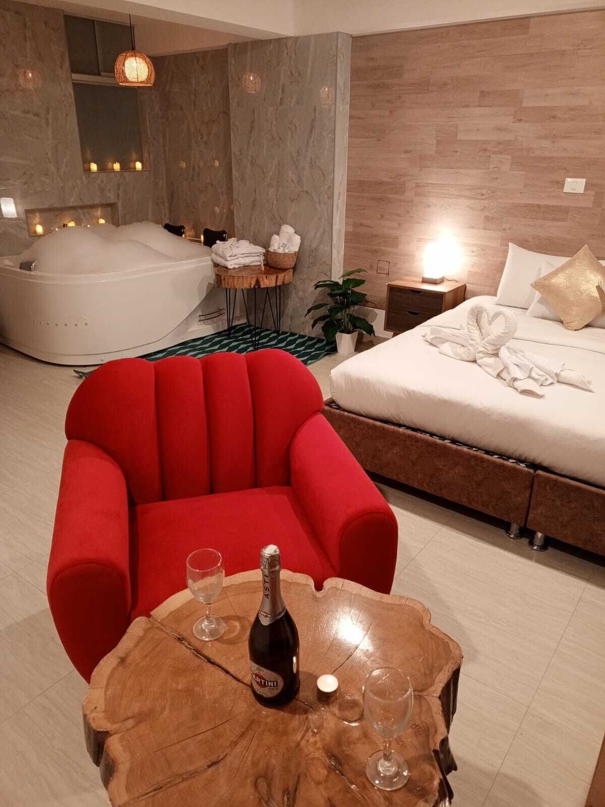 Una habitación de hotel moderna con un sillón rojo, una cama grande, un jacuzzi y una mesa de madera con una botella de Martini y copas.
