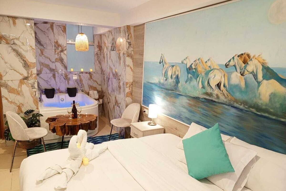 A hotel room with a large bed, a painting of horses, a jacuzzi, and a dining area with a wooden table and chairs.