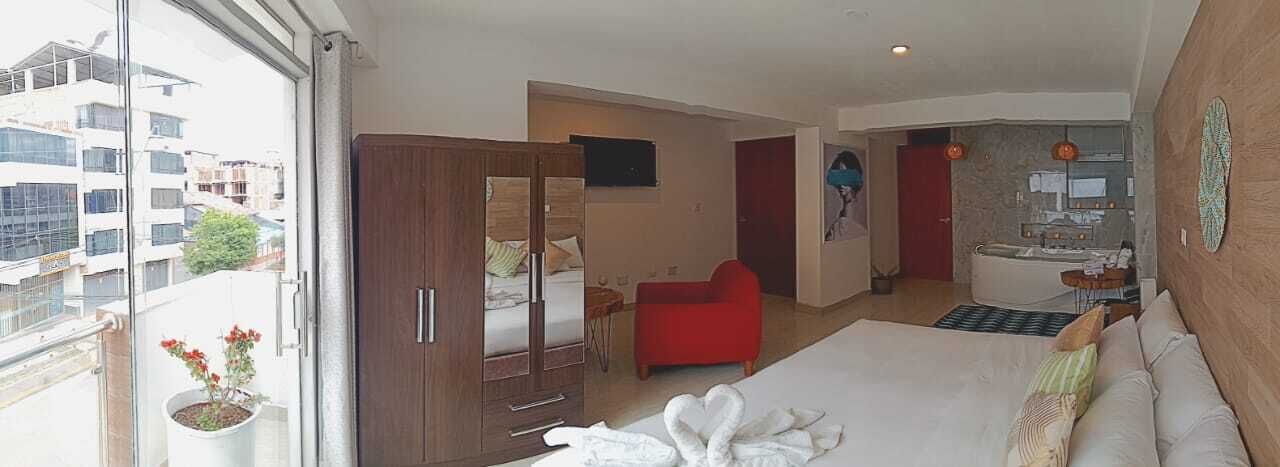 Habitación de hotel con cama, un sillón rojo, un armario de madera, baño con bañera y vistas a la ciudad a través de un ventanal grande.