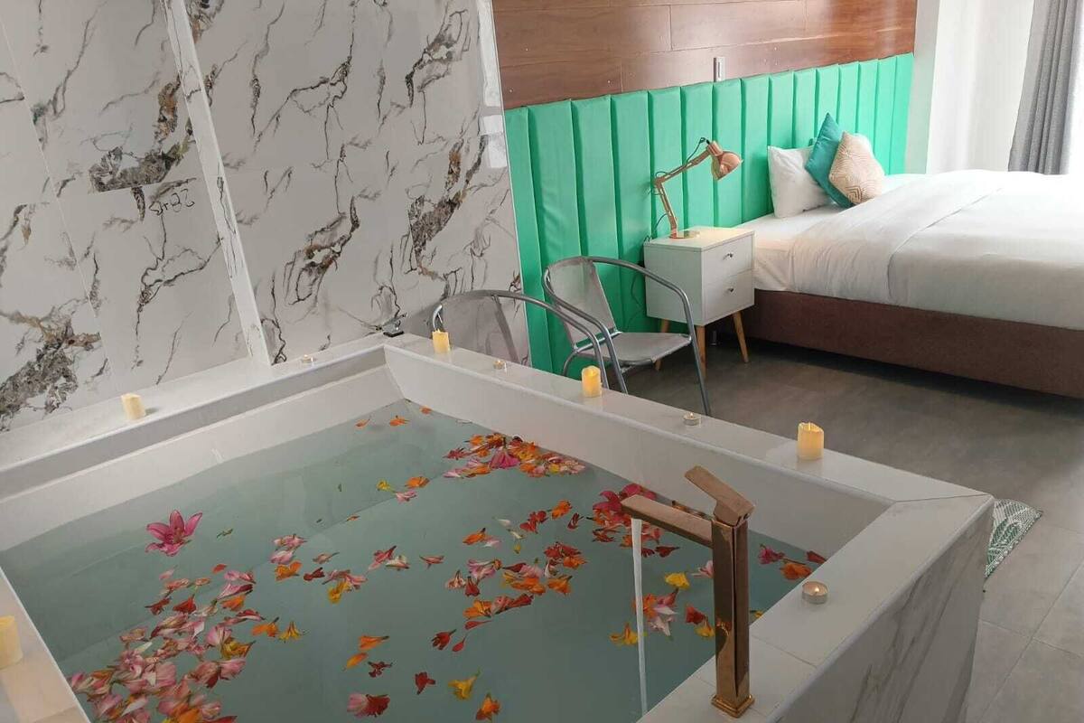 A bathroom with a marble vanity, a bathtub with floating flower petals, a green upholstered headboard, and a bedside table with a lamp.