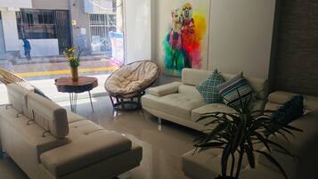 Un salón moderno con un sofá blanco, una silla de ratán y una mesa de centro. Hay un ventanal con vistas a la calle y un cuadro colorido en la pared.