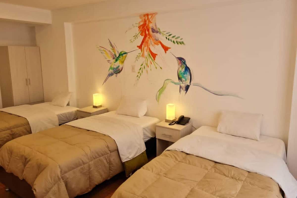 A hotel room with two beds, each with a beige duvet and white pillows. There are bedside tables with lamps and a phone. The walls feature a mural of colorful birds.