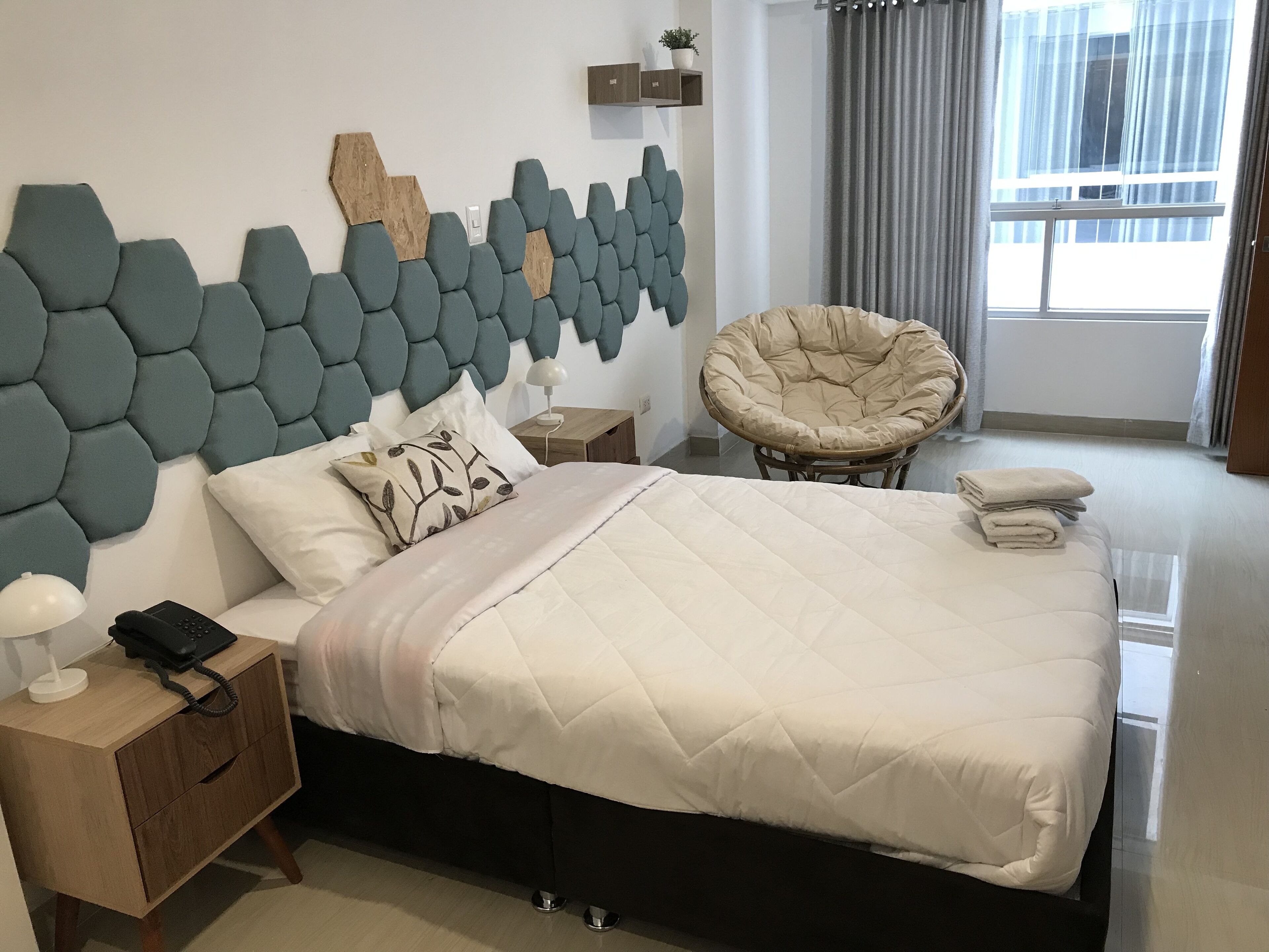 A modern bedroom with a large bed, a headboard with a geometric pattern, a bedside table with a lamp, a chair, and a window with curtains.