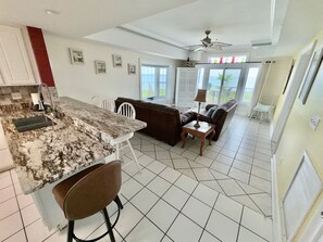 A kitchen with a marble countertop, white chairs, a brown sofa, a small table with a lamp, and a ceiling fan. - Ship-Watch Dunes ~ Oceanfront ~ Dolphins! Pools! (Tybee Island)