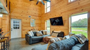 Un intérieur de chalet chaleureux avec un canapé d’angle, un téléviseur à écran plat fixé au mur et de grandes fenêtres offrant une vue sur l’extérieur.
