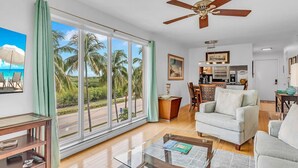 A spacious living room with a large sliding glass door leading to a balcony with a view of palm trees and the ocean. There is a ceiling fan, a dining area with a table and chairs, and a kitchen in the background. - Updated Unit with Beautiful Florida Bay Sunsets (Islamorada)