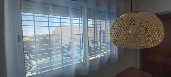 A room with a large window with horizontal blinds, a woven light fixture, and a view of a building outside.