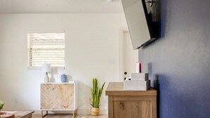 Une pièce comprenant un meuble avec un motif en chevrons, un buffet en bois, un téléviseur fixé au mur, une fenêtre avec des stores et une plante en pot.