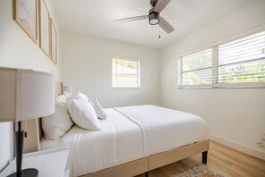 Una camera da letto con un letto, una ventola a soffitto, due finestre e un comodino con una lampada.