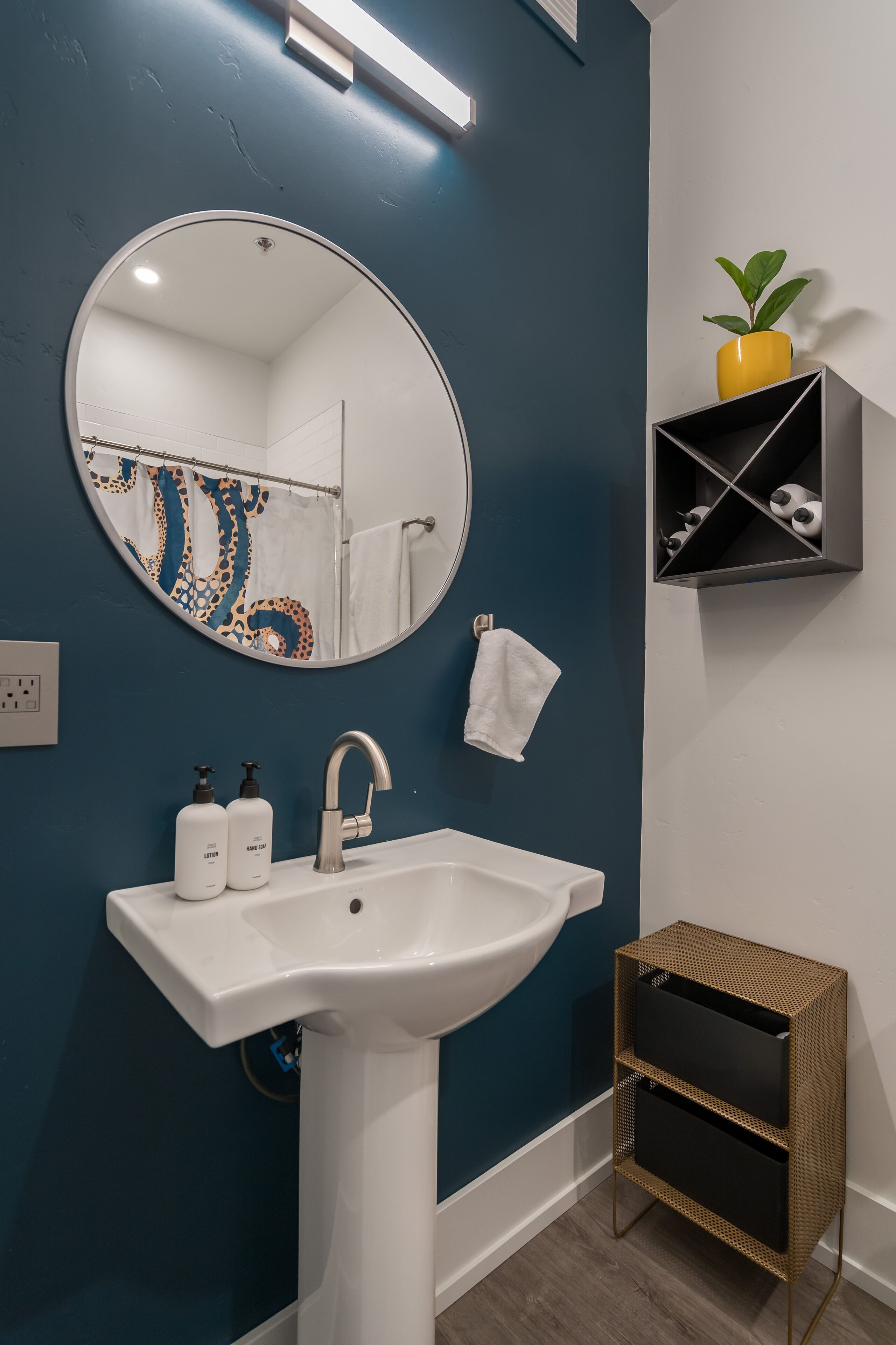 A bathroom with a white sink, a round mirror, a towel rack, and a shelf with a plant.