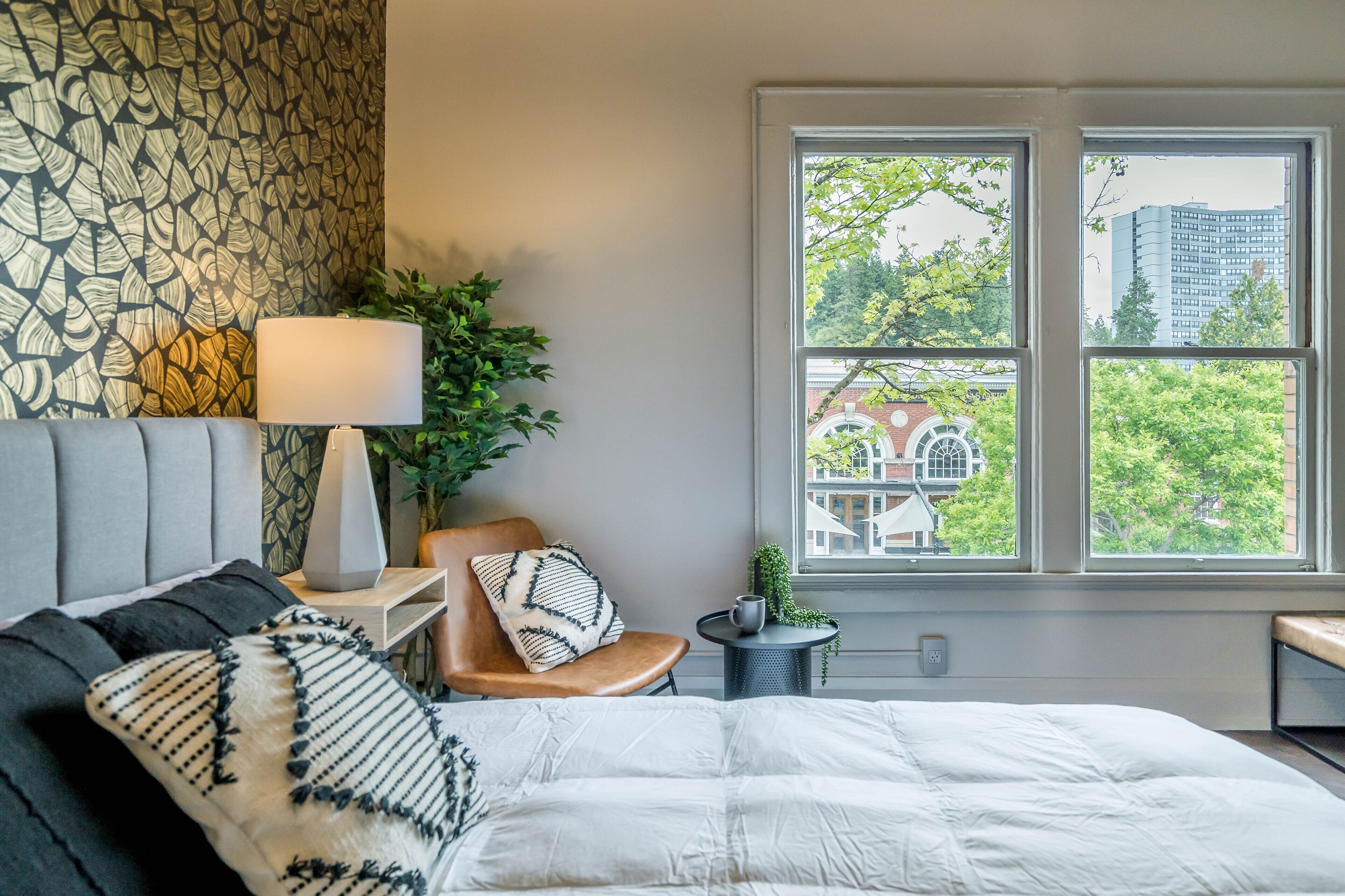 Un dormitorio con una cama grande, una silla, una mesita de noche con una lámpara y una ventana con vistas a edificios y árboles.