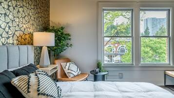 Un dormitorio con una cama grande, una silla, una mesita de noche con una lámpara y una ventana con vistas a edificios y árboles.
