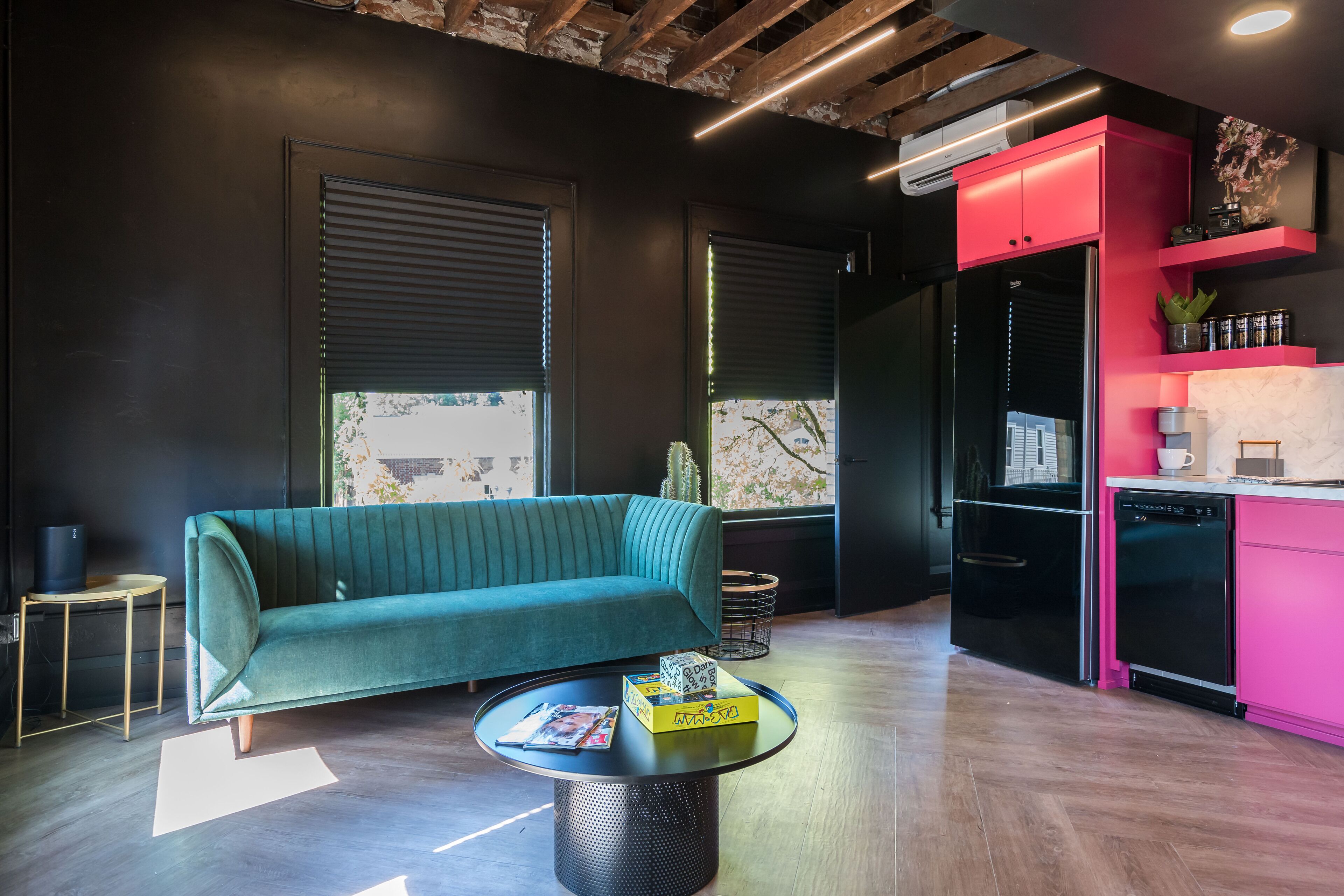 A modern living room with a teal sofa, a round coffee table with magazines, and a kitchen area with pink cabinets and black appliances.