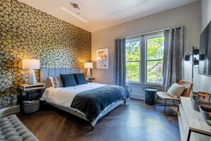 A modern bedroom with a patterned wallpaper, a large bed, bedside lamps, a chair, a desk, and a window with curtains. - The 505 Eugene (Eugene)