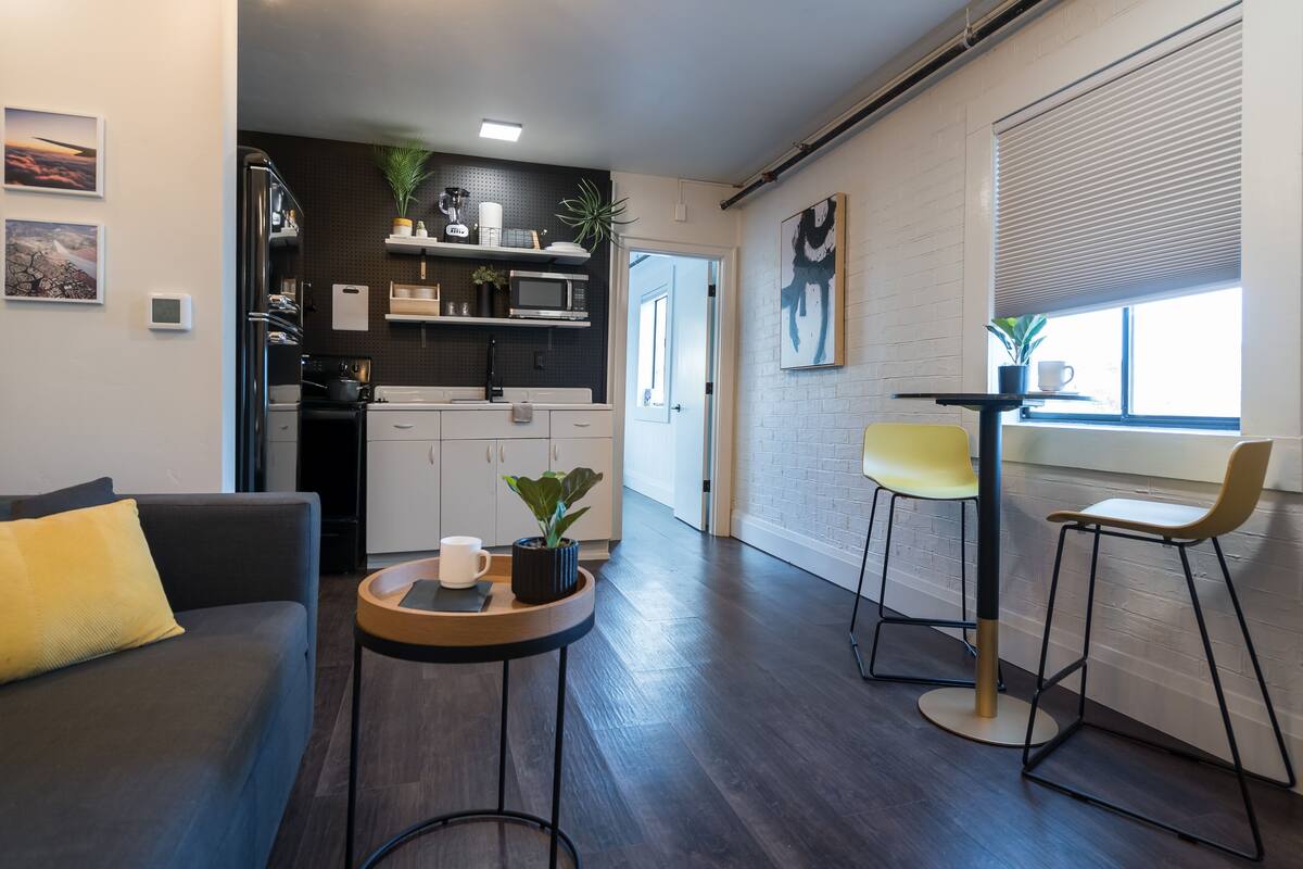 A modern living room with a sofa, a coffee table, and a dining area with chairs. There is a kitchen in the background with appliances and plants.