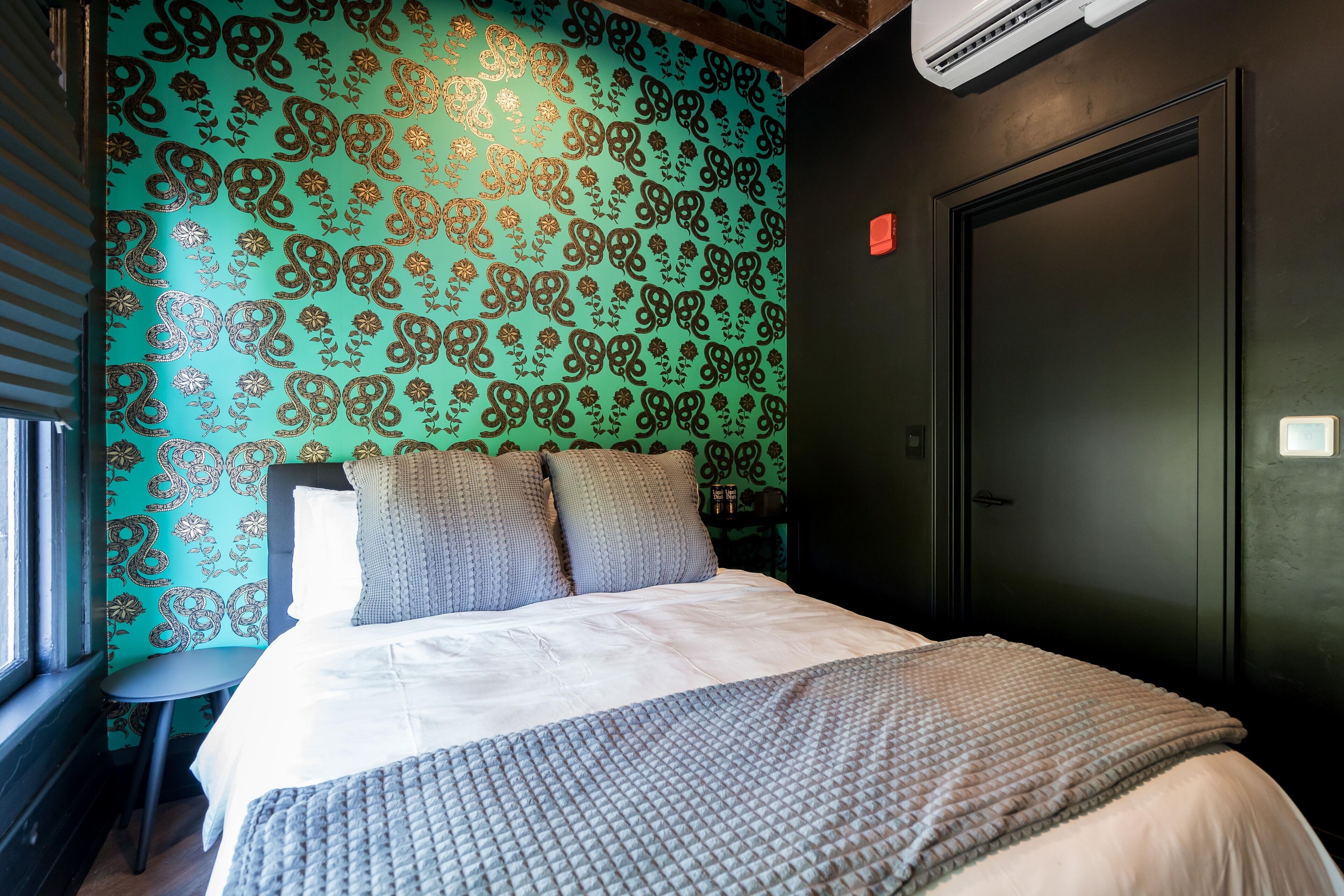Un lit bien fait, avec une couverture et des oreillers gris, une fenêtre avec des stores et un mur orné d’un motif cachemire.