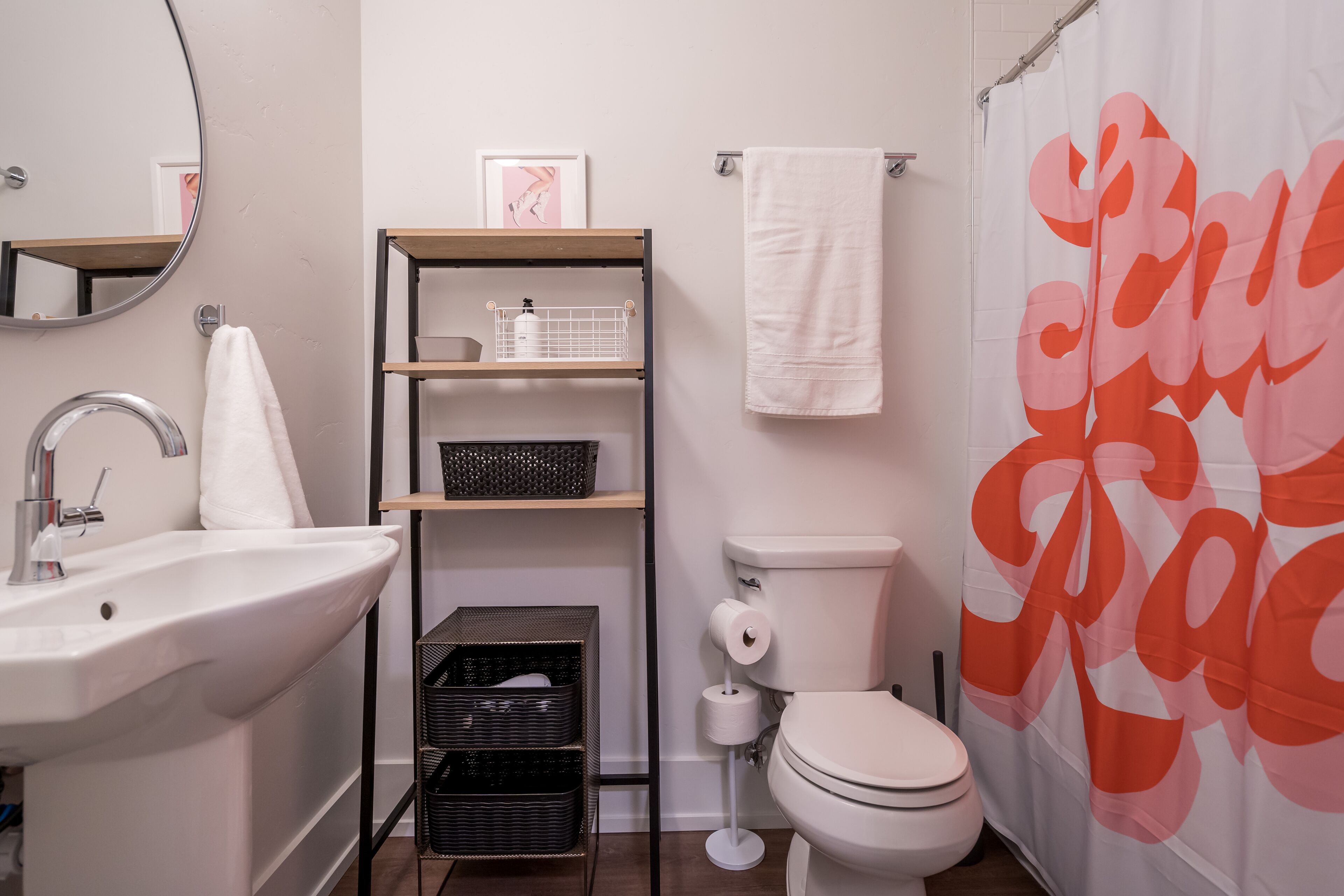 Un baño con lavamanos, inodoro y cortina de ducha blanca, que presenta un logotipo grande. Hay estantes con cestas y un cuadro en la pared.