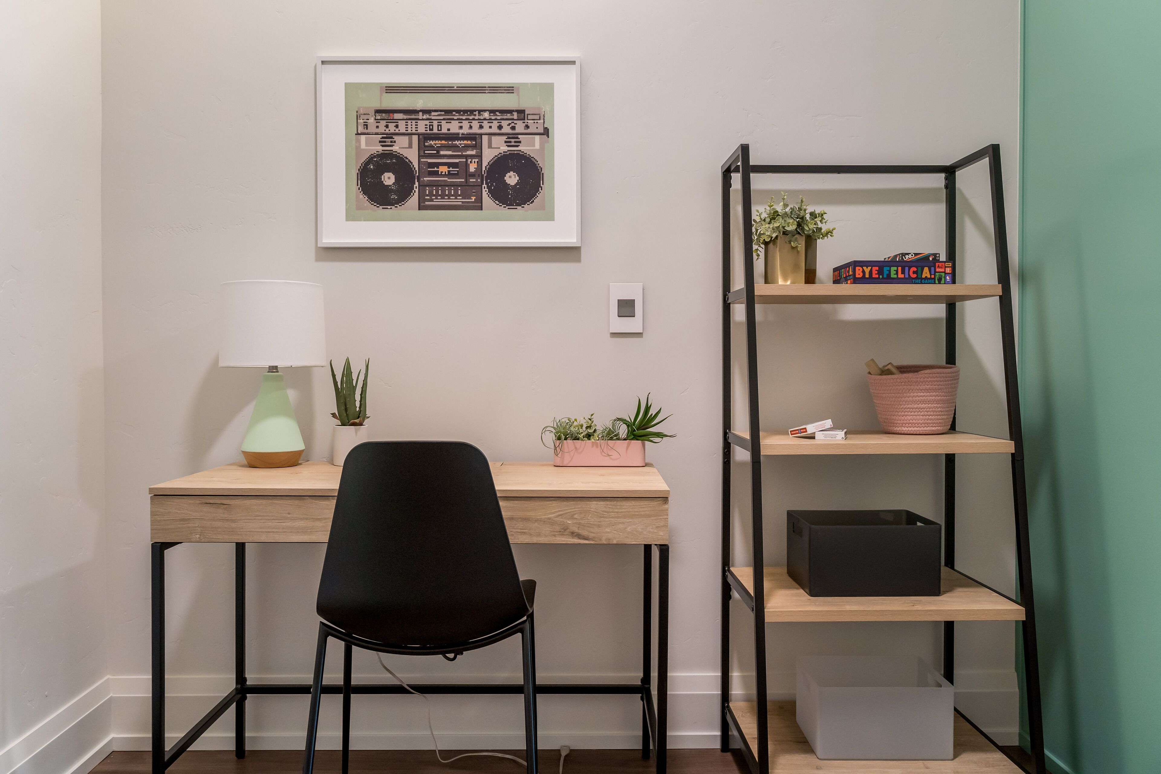 Un bureau avec une chaise, un tableau encadré, une lampe et une étagère avec des plantes et une boîte.