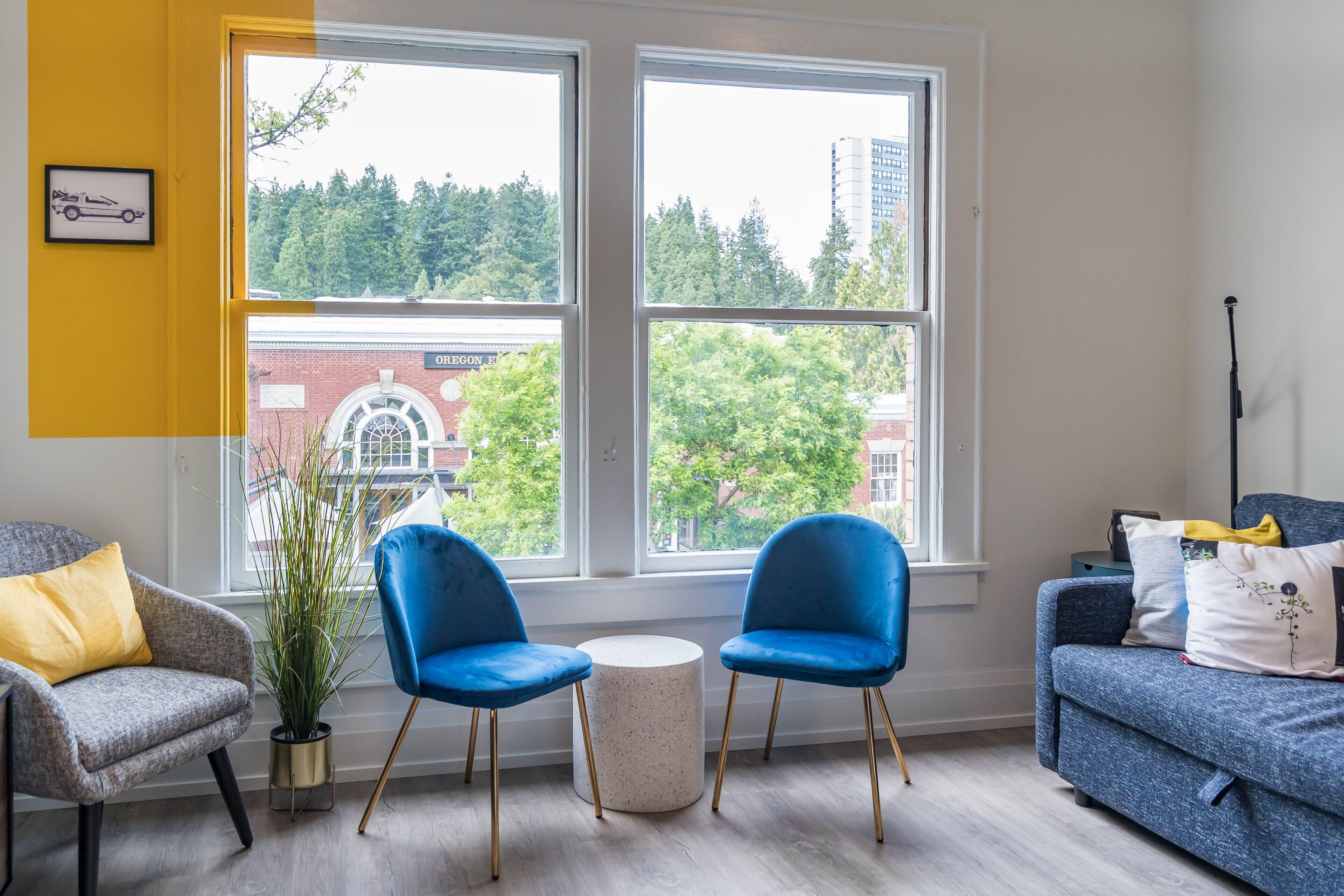 Une pièce avec deux chaises bleues, un fauteuil gris et un canapé bleu. Une petite table ronde se trouve au centre. De grandes fenêtres offrent une vue sur les arbres et les bâtiments à l’extérieur.