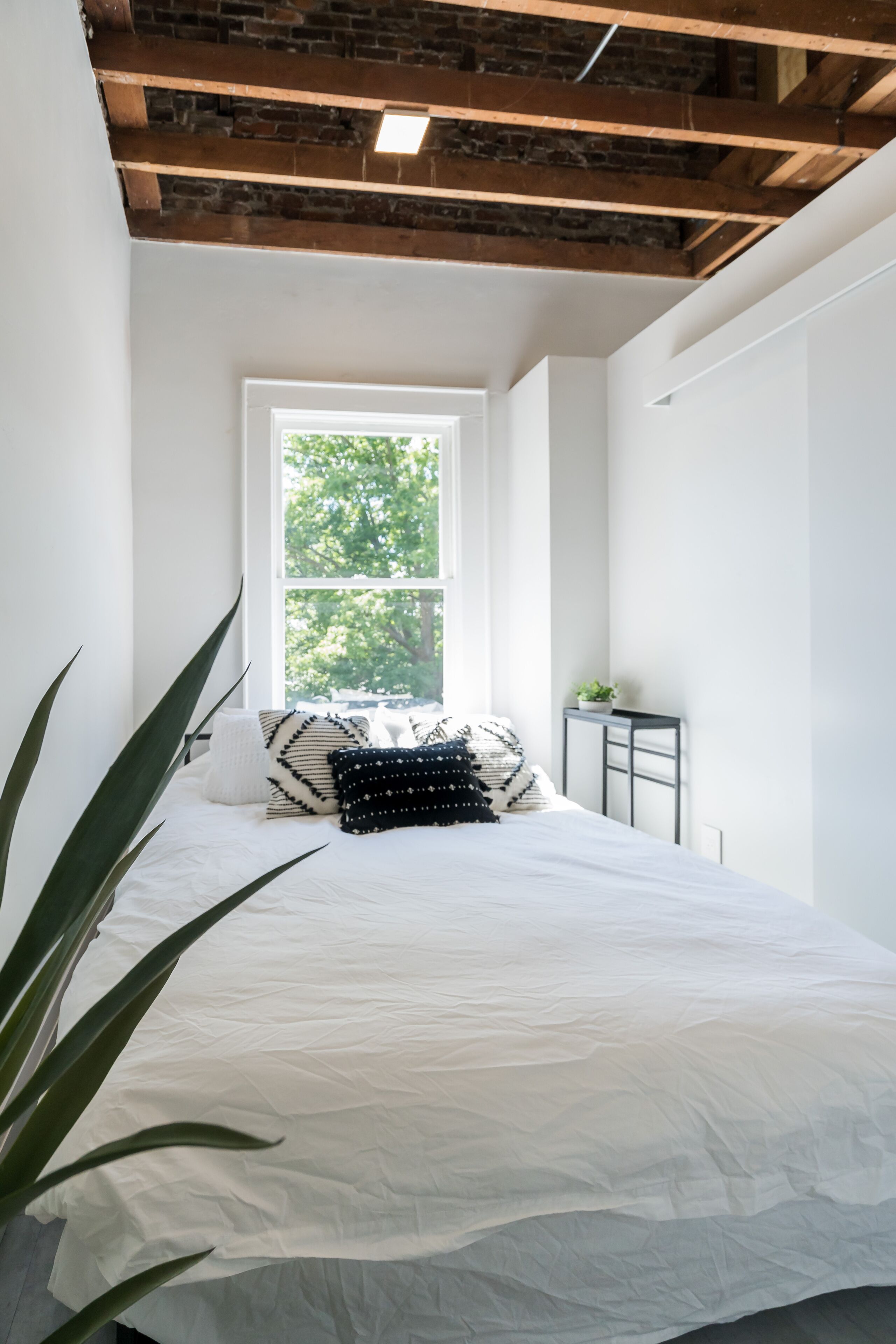 A bedroom with a large window, a bed with white bedding, a small table with a plant, and a brick ceiling.