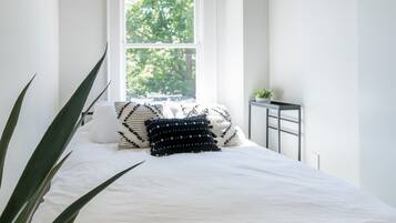 A bedroom with a large window, a bed with white bedding, a small table with a plant, and a brick ceiling.